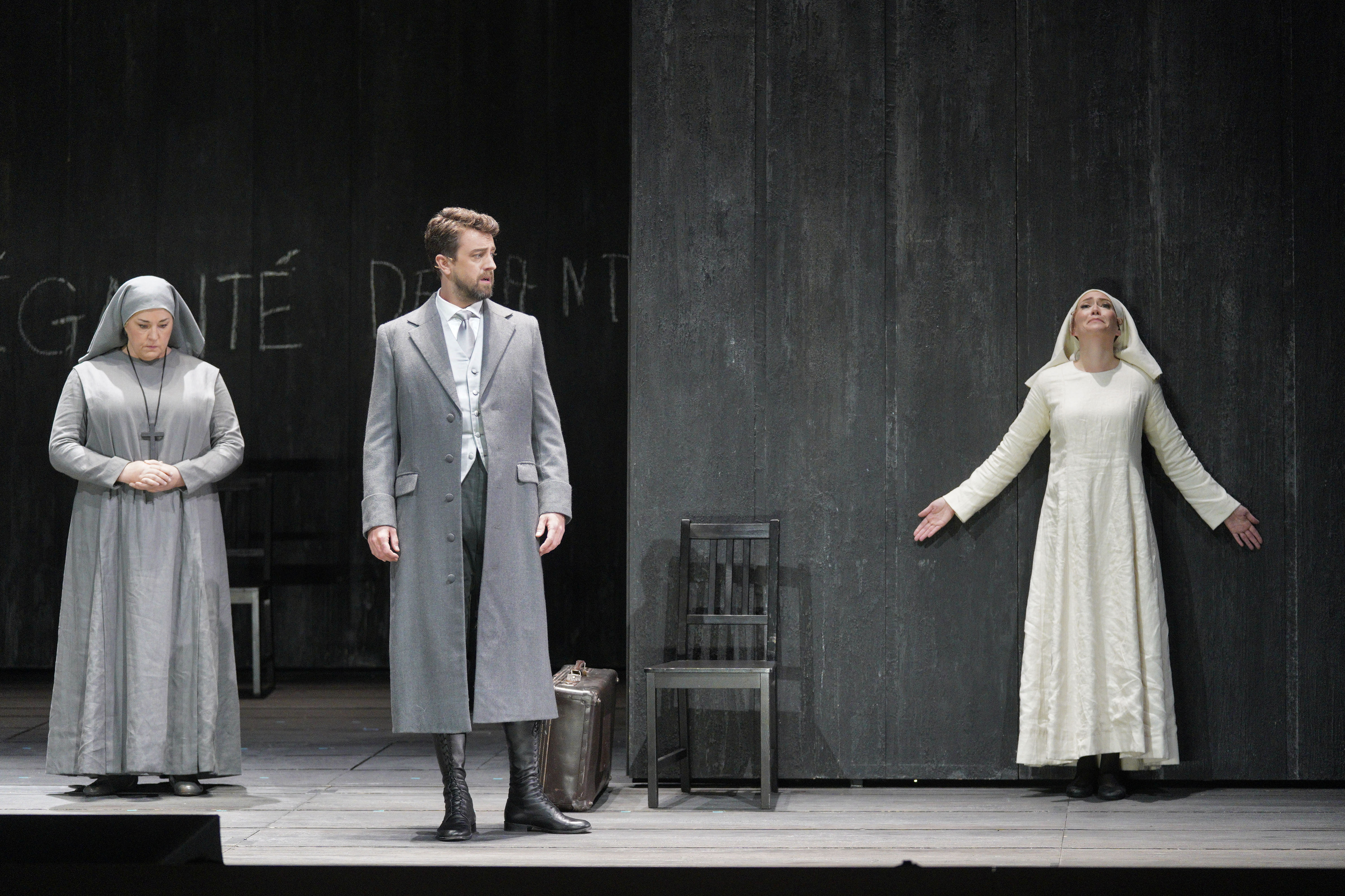 A nun and a man in a gray coat stand apart from a woman in white against a dark stage wall.