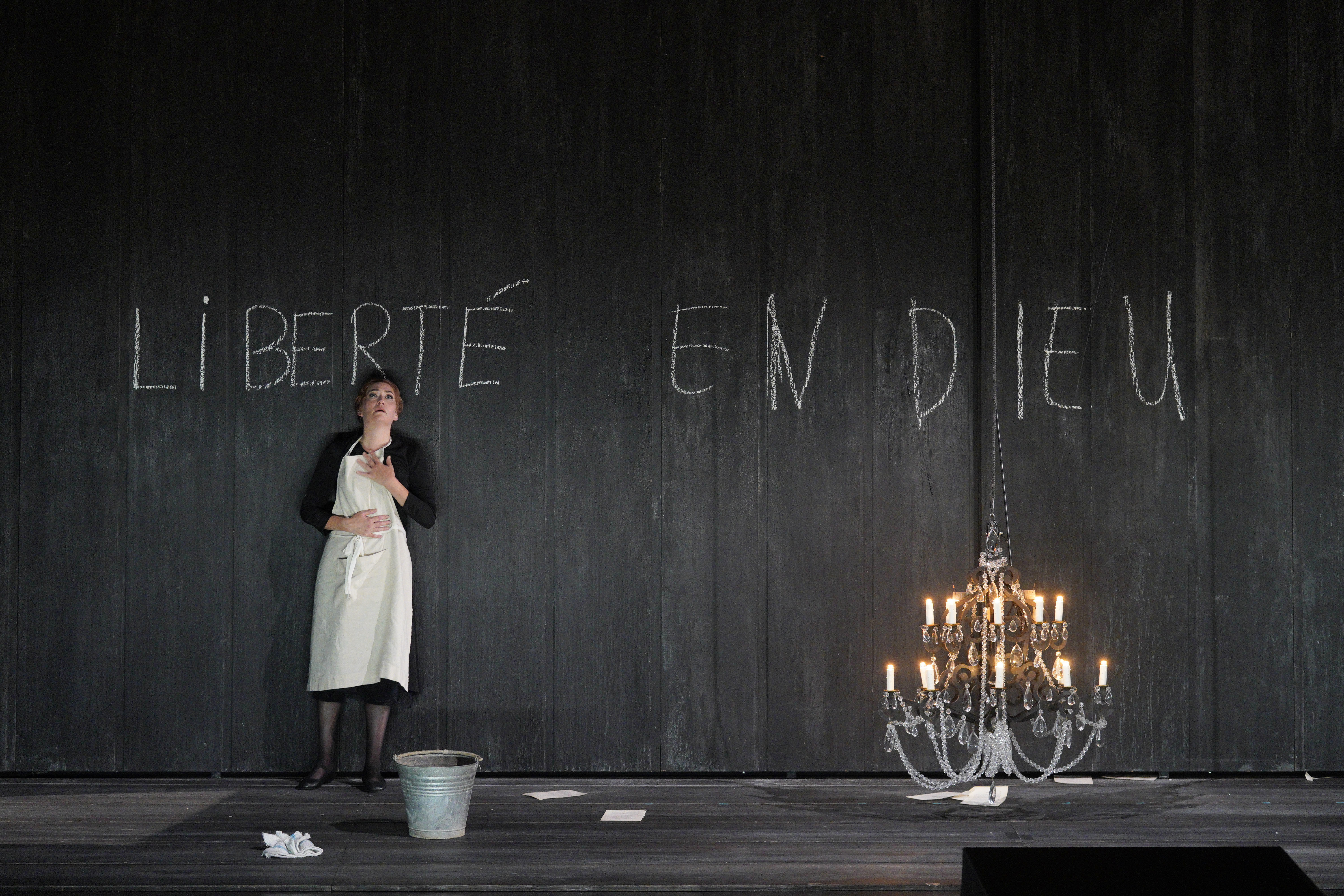 Blanche, wearing a white apron, stands against a black wall chalked with “Liberté en Dieu,” beside a lit chandelier.